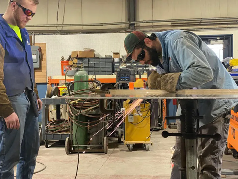 Two men working in a workshop. One man is welding metal on a table, creating sparks, while the other watches.